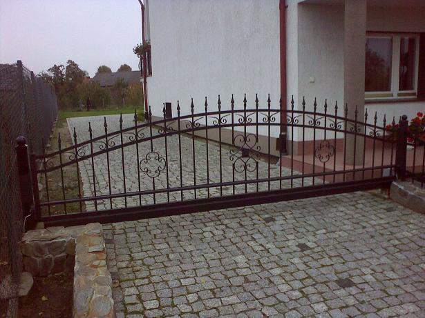 Przesuwna, metalowa brama wjazdowa z kutymi zdobieniami w kształcie serc i rozet, zamontowana na posesji z brukowaną kostką i ogrodzeniem panelowym w tle.