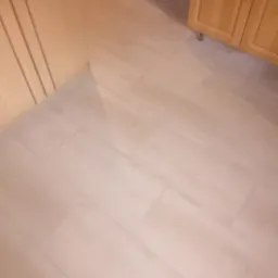 Jasne, drewnopodobne płytki podłogowe ułożone w jodełkę w kuchni, widoczne fragmenty zabudowy kuchennej z jasnego drewna.