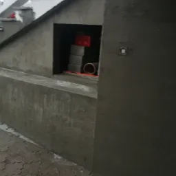 Surowa ściana fundamentowa z wnęką, w której składowane są bloczki betonowe i rura kanalizacyjna, widoczny poziomica, szare wykończenie powierzchni, kostka brukowa na chodniku.