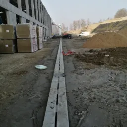 Budowa hali: widok na długi betonowy odpływ liniowy, palety materiał&oacute;w izolacyjnych w folii, w tle ściany budynku w stanie surowym otwartym, kopiec ziemi i pojazdy budowlane.