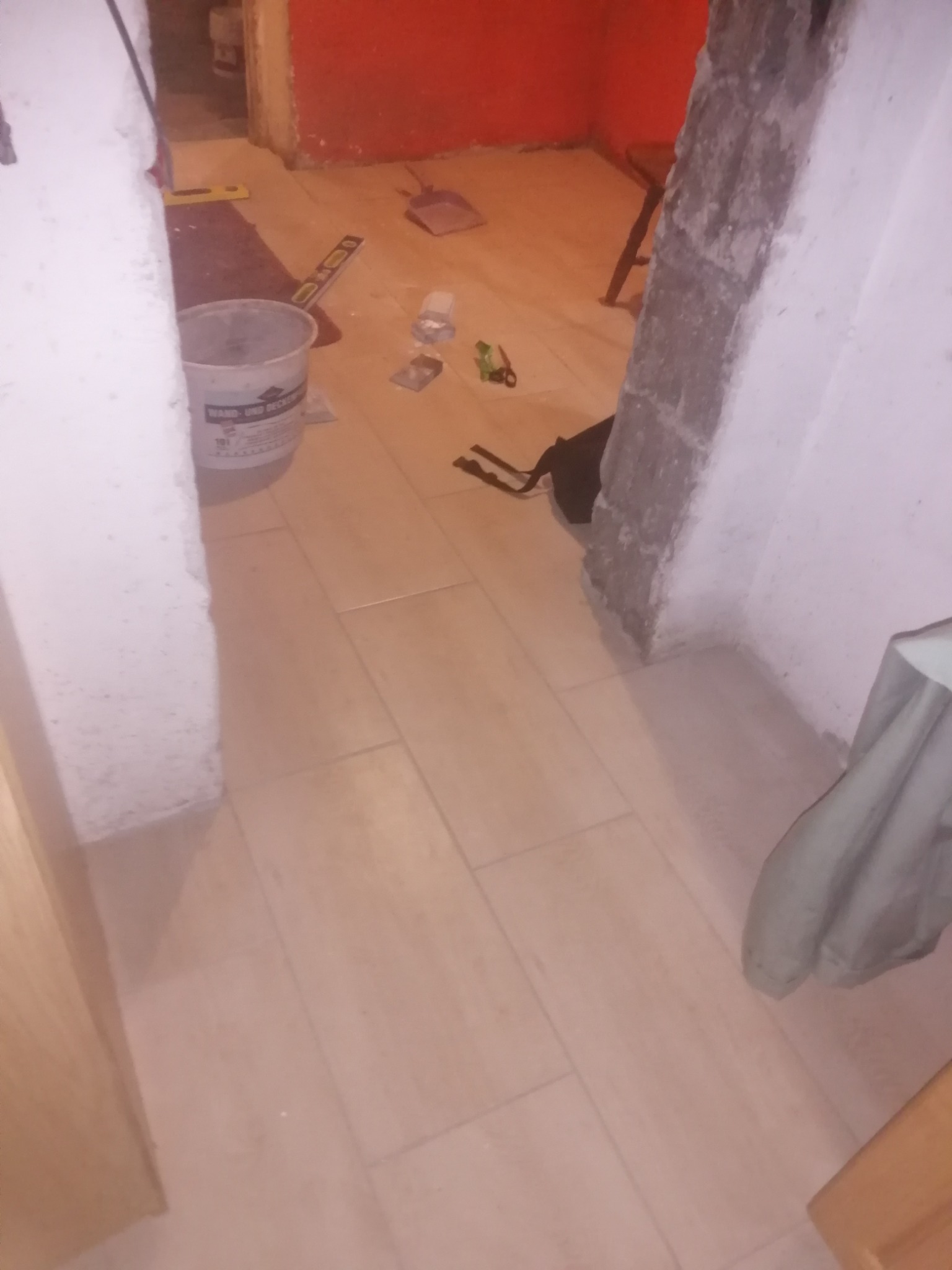 Podłoga wyłożona jasnymi płytkami, widoczny fragment ściany z surowym tynkiem, wiadro z zaprawą, poziomica i narzędzia do układania płytek w trakcie remontu.