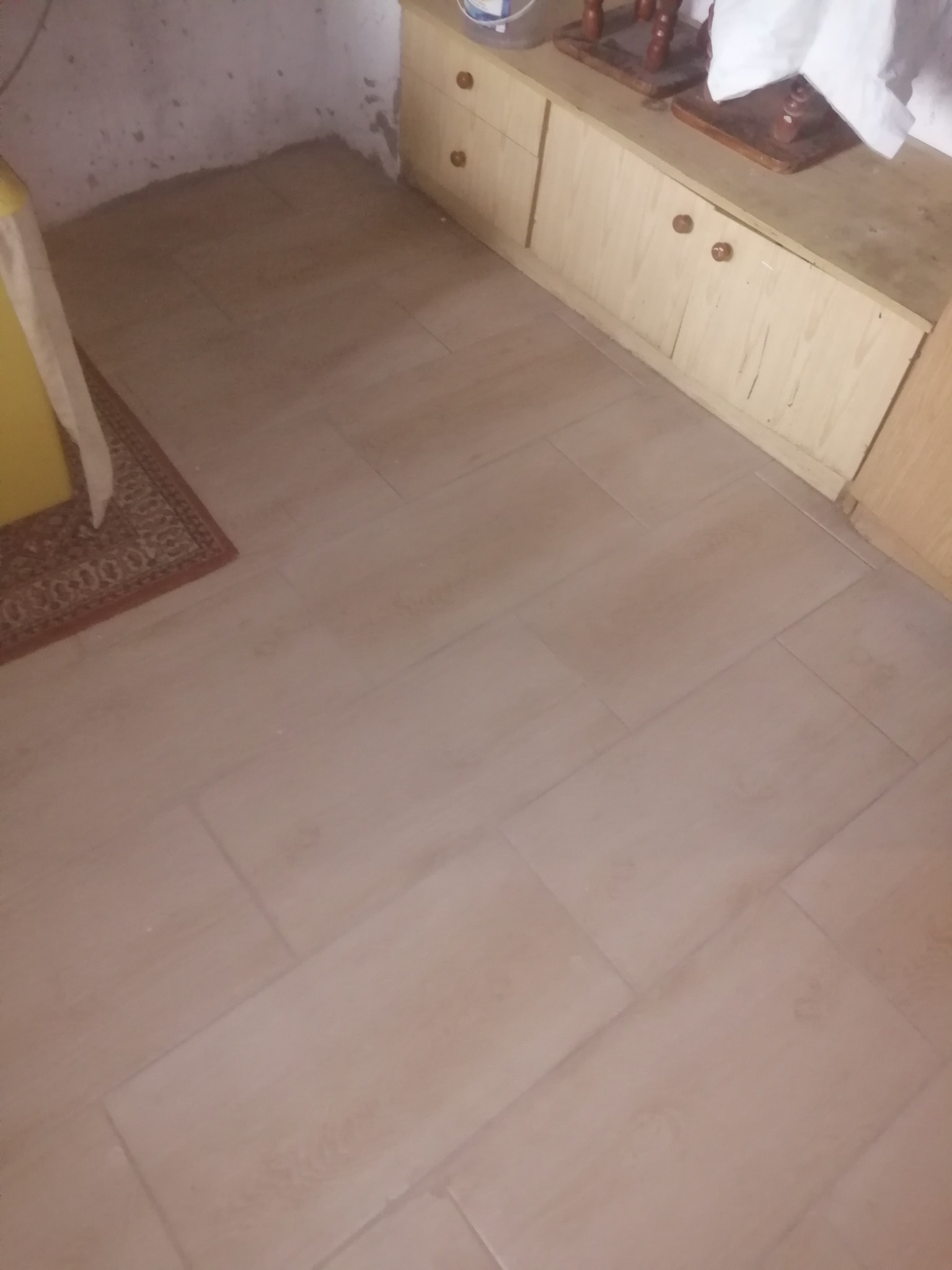 Jasne, prostokątne płytki podłogowe ułożone w pomieszczeniu z fragmentem szafki z jasnego drewna i dywanikiem z brązowymi zdobieniami.