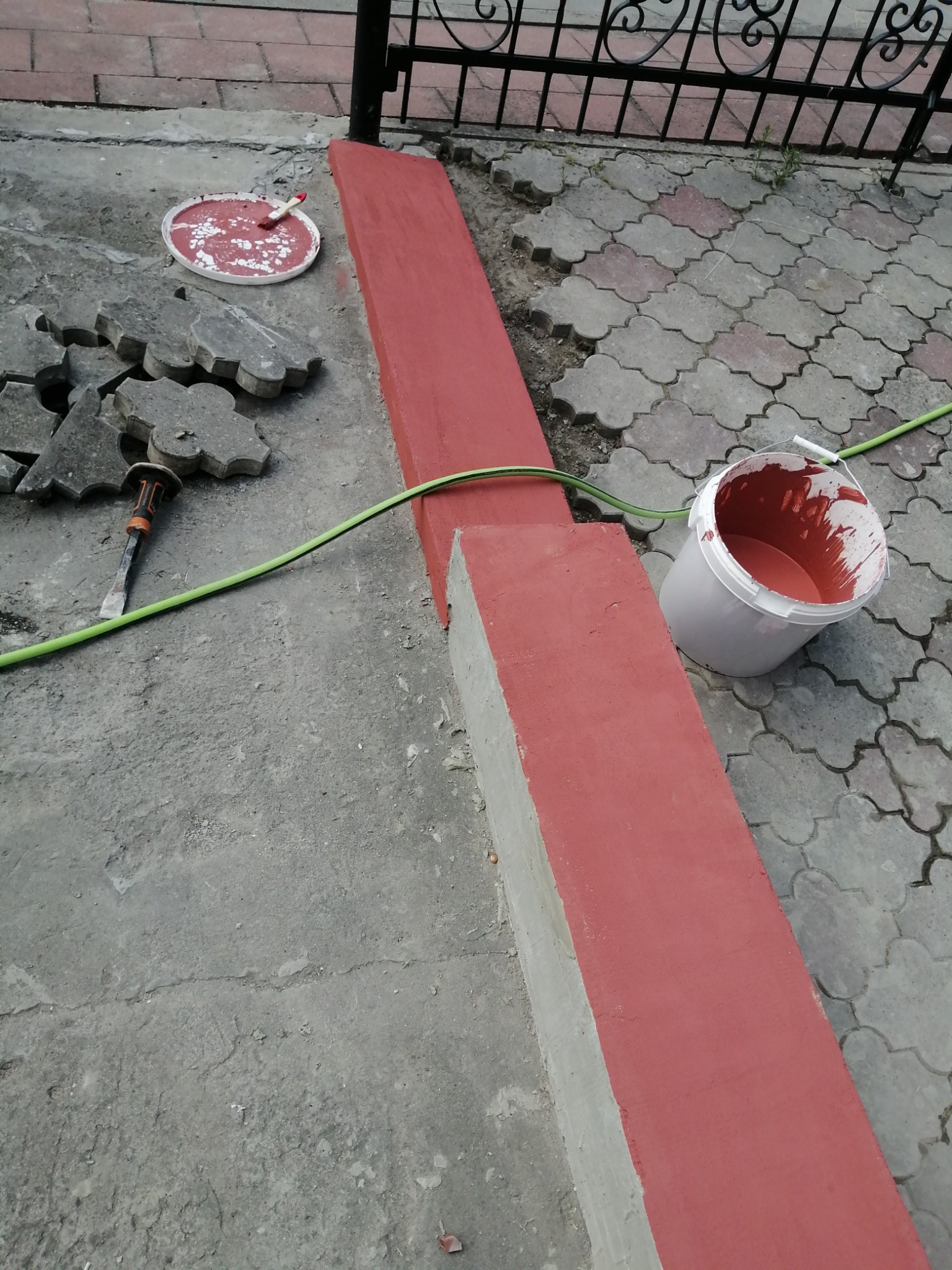 Świeżo pomalowany, ceglastoczerwony cokół budynku z widocznymi śladami farby, obok wiadro z farbą, pędzel w kuwecie, dłuto i kostka brukowa na betonowym podłożu.