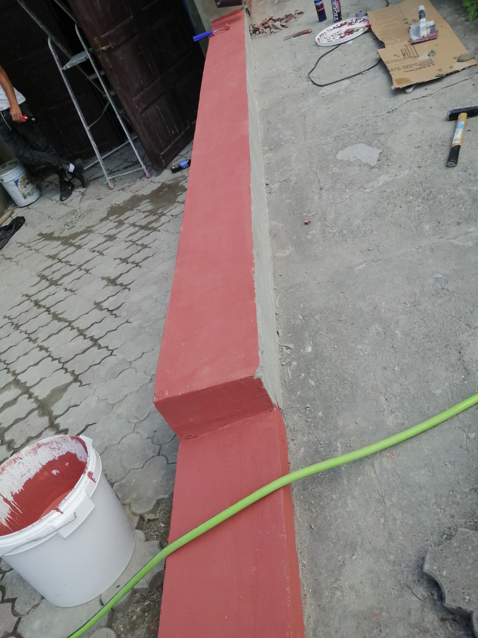 Świeżo pomalowany na czerwono betonowy cokół budynku, widoczny biały kubeł z farbą, narzędzia malarskie i zielony wąż ogrodowy na szarym chodniku.