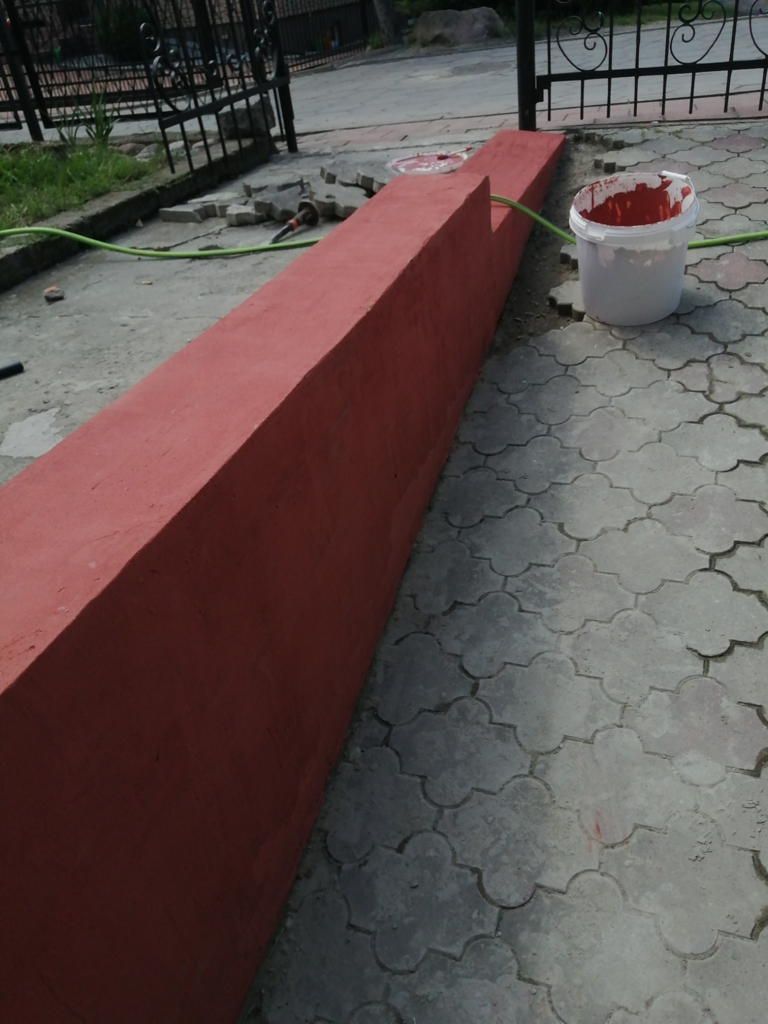 Świeżo pomalowany na czerwono betonowy murek przy posesji, obok wiadro z farbą i kostka brukowa, w tle metalowe ogrodzenie.