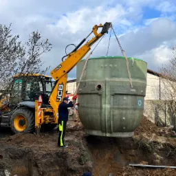 Montaż przydomowej oczyszczalni ścieków w Czernikowie: Żółta koparko-ładowarka JCB opuszcza zbiornik do wykopu, pracownik w stroju roboczym kontroluje proces.