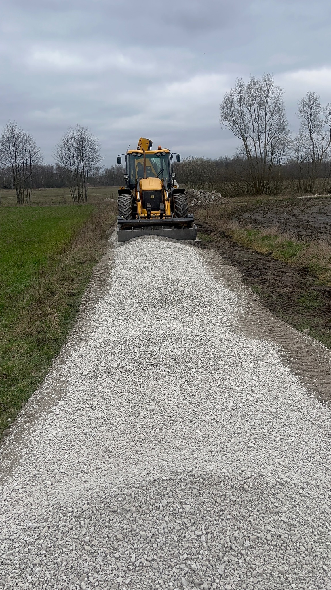 Żółta koparko-ładowarka rozścieła warstwę jasnego kruszywa na ziemnej drodze, widok z przodu na tle pochmurnego nieba i nagich drzew.