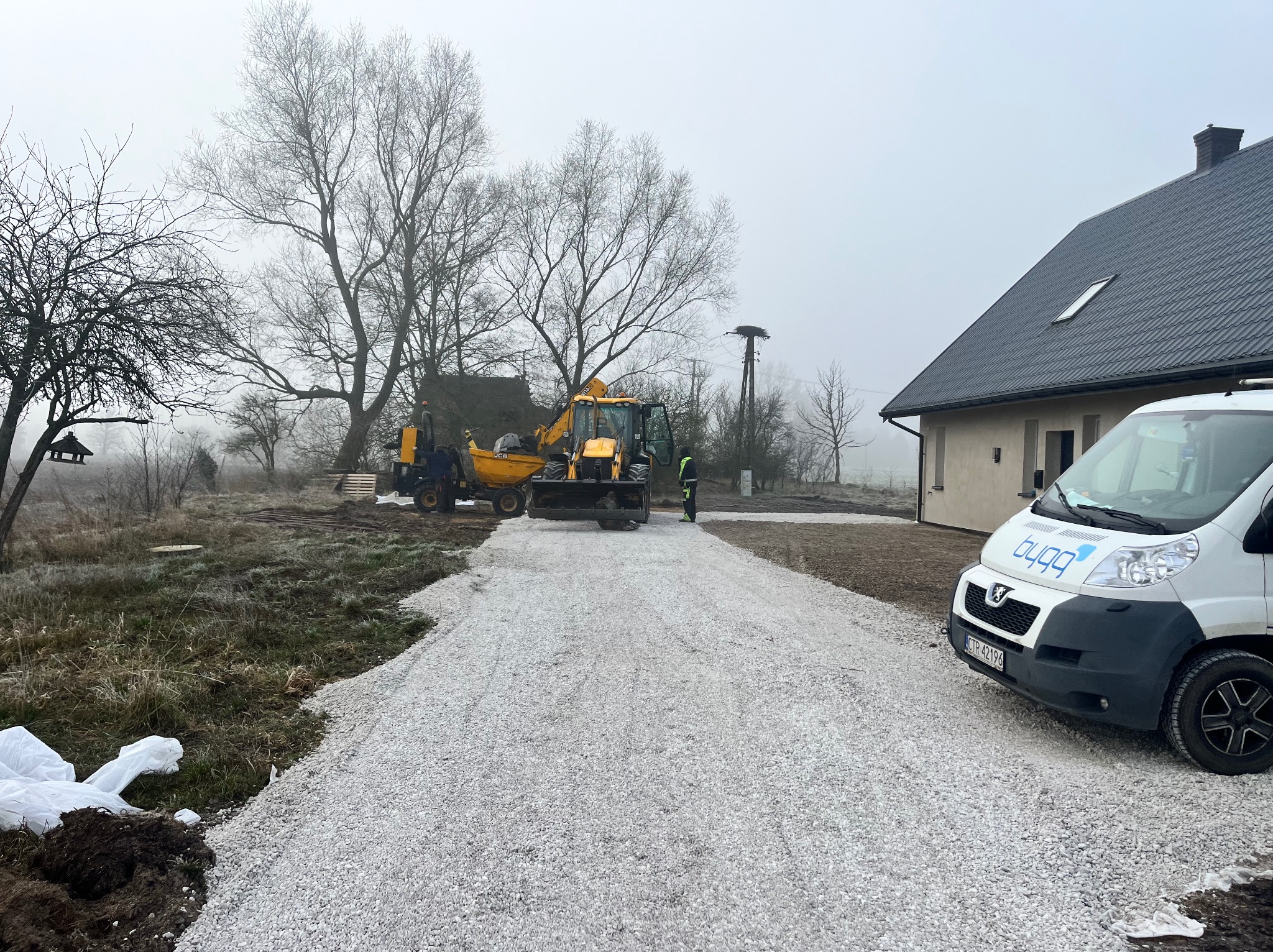 Żwirowa droga dojazdowa w trakcie budowy, z widocznym sprzętem budowlanym i samochodem dostawczym firmy, pochmurny dzień.