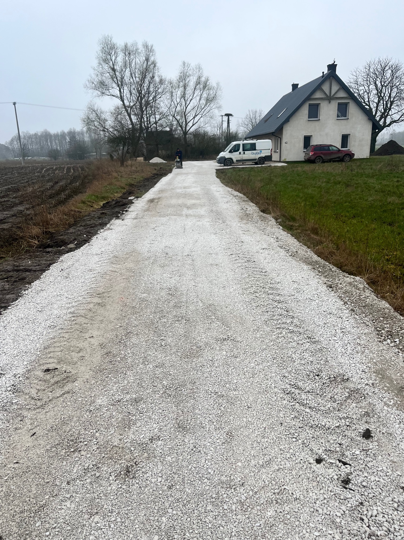 Nowo utwardzona droga dojazdowa wysypana jasnym żwirem, widoczny pracownik z maszyną, w tle dom jednorodzinny i zaparkowane pojazdy w pochmurny dzień.