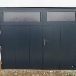 Dwuskrzydłowa, grafitowa brama garażowa z dwoma poziomymi oknami z matowego szkła, zamontowana w białej ścianie budynku. Na dole widoczne ziemia i fragment ceglanej podmurówki.