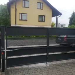 Nowoczesna, szara brama wjazdowa z perforowanymi panelami i automatyką, na tle domu z żółtą elewacją i szarą kostką brukową.