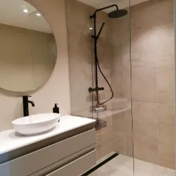 Nowoczesna łazienka z beżowymi płytkami, kabiną prysznicową typu walk-in z czarną armaturą i okrągłym lustrem nad umywalką nablatową.