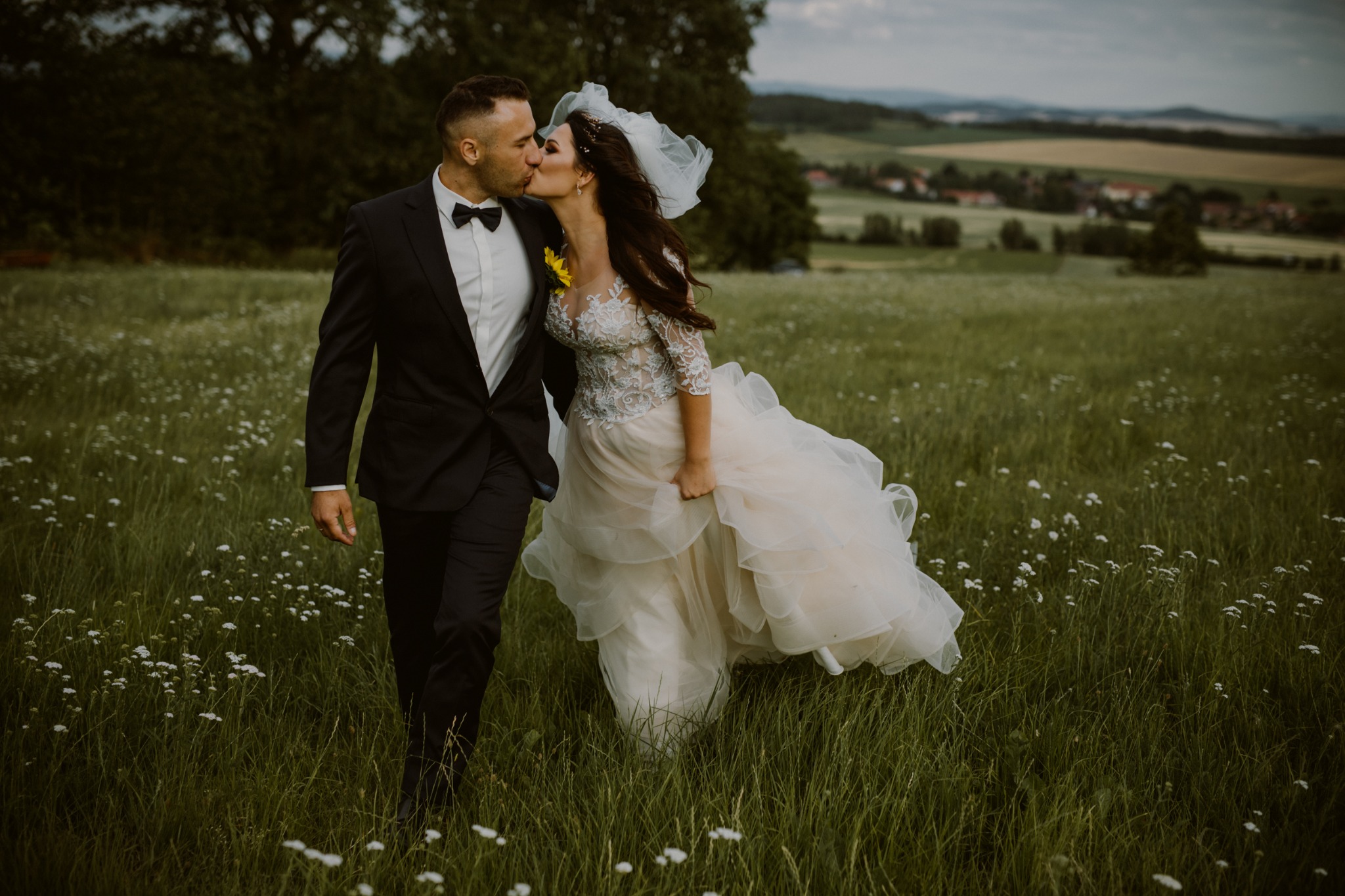 Para młoda w czarnym garniturze i koronkowej sukni ślubnej z welonem, całująca się na łące z białymi kwiatami, w tle zielone wzgórza i zabudowania.