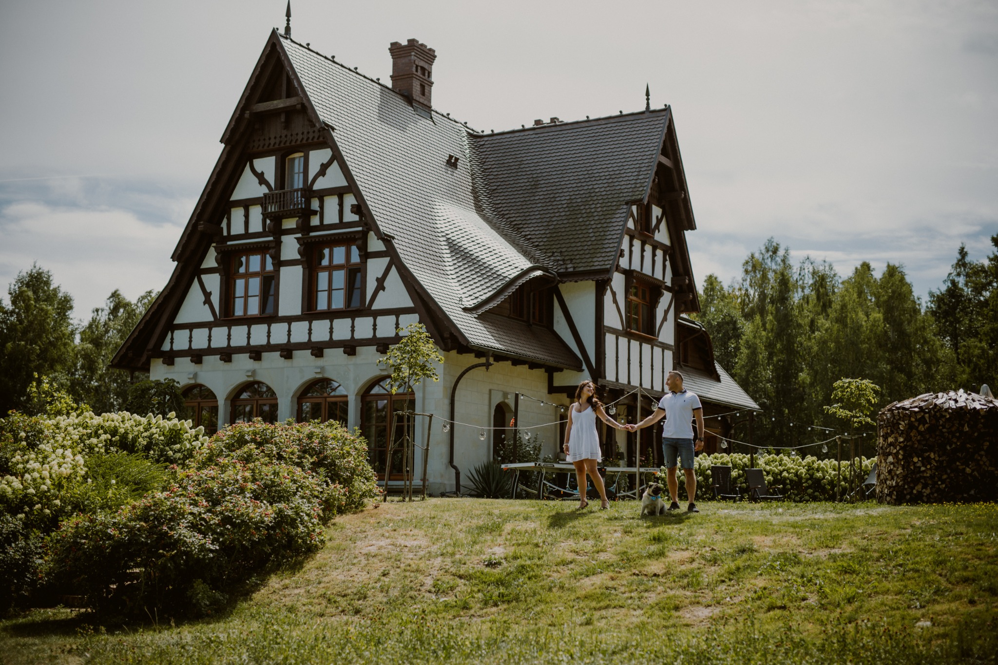 Para trzymająca się za ręce spaceruje z psem rasy mops przed domem w stylu szachulcowym z ogrodem, w tle stos drewna.
