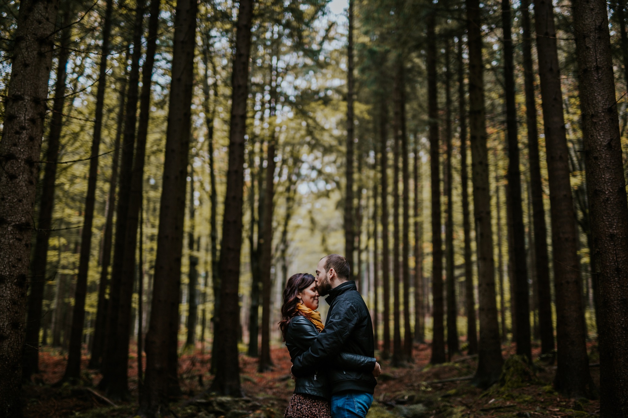 Zakochana para w czarnych kurtkach obejmuje się w lesie, mężczyzna całuje kobietę w czoło, wysokie drzewa w tle tworzą tunel.