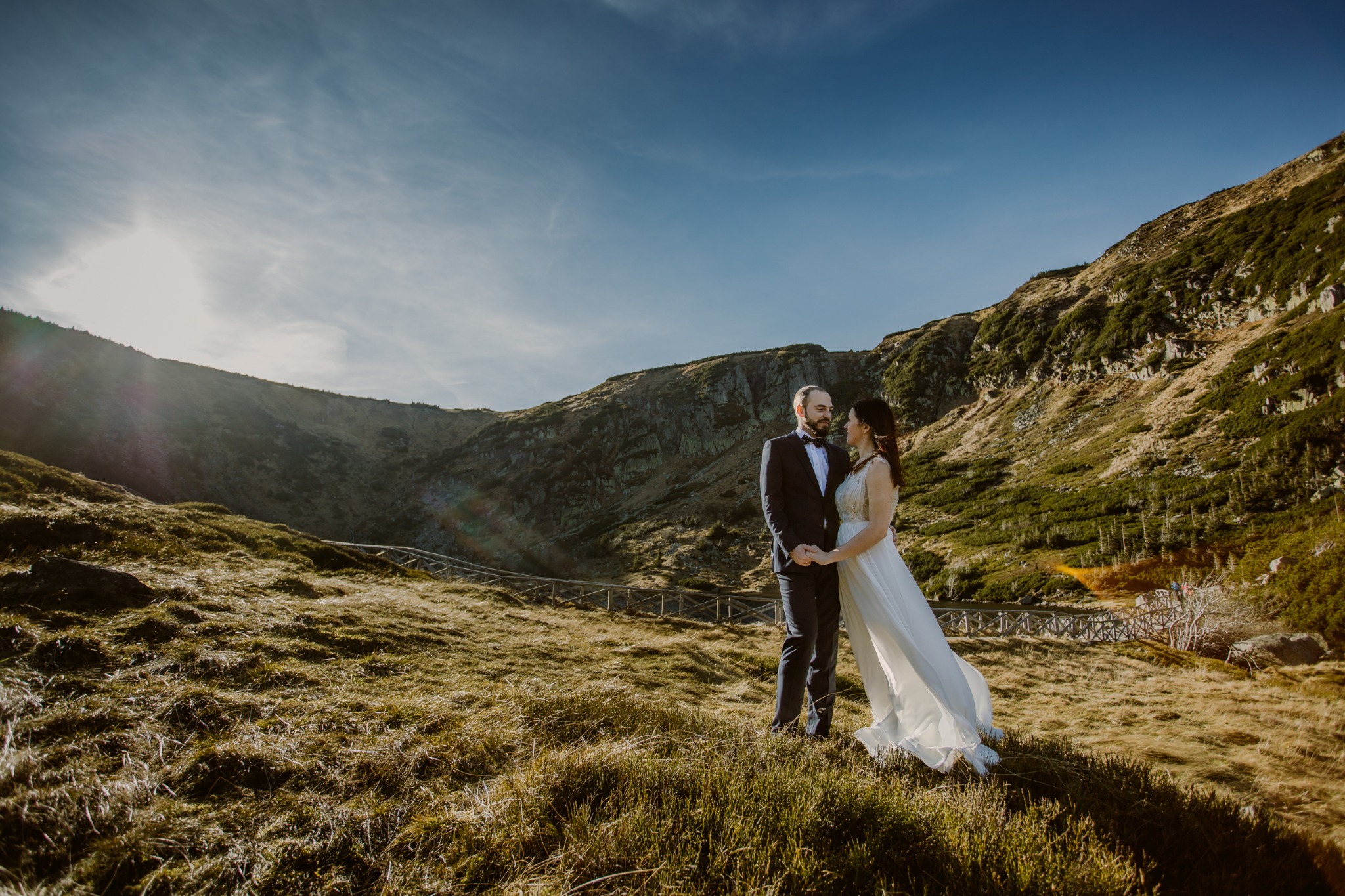 Para młoda trzymająca się za ręce na tle górskiego krajobrazu z drewnianym mostkiem w oddali, oświetlona promieniami słońca.