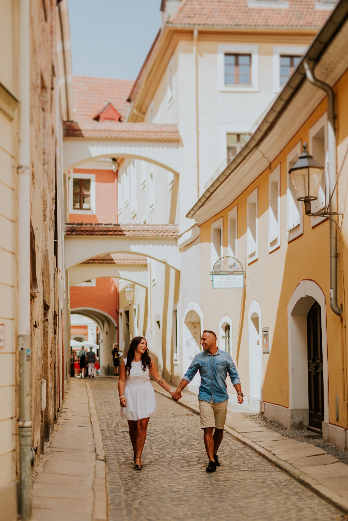 Uśmiechnięta para trzymająca się za ręce spaceruje brukowaną uliczką między kamienicami w słoneczny dzień.
