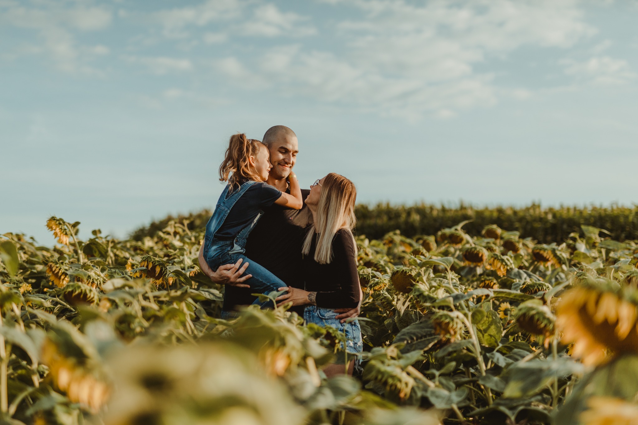 Rodzina w polu słoneczników: tata trzyma córkę w dżinsowych ogrodniczkach, mama patrzy na nich z uśmiechem, ciepłe światło.