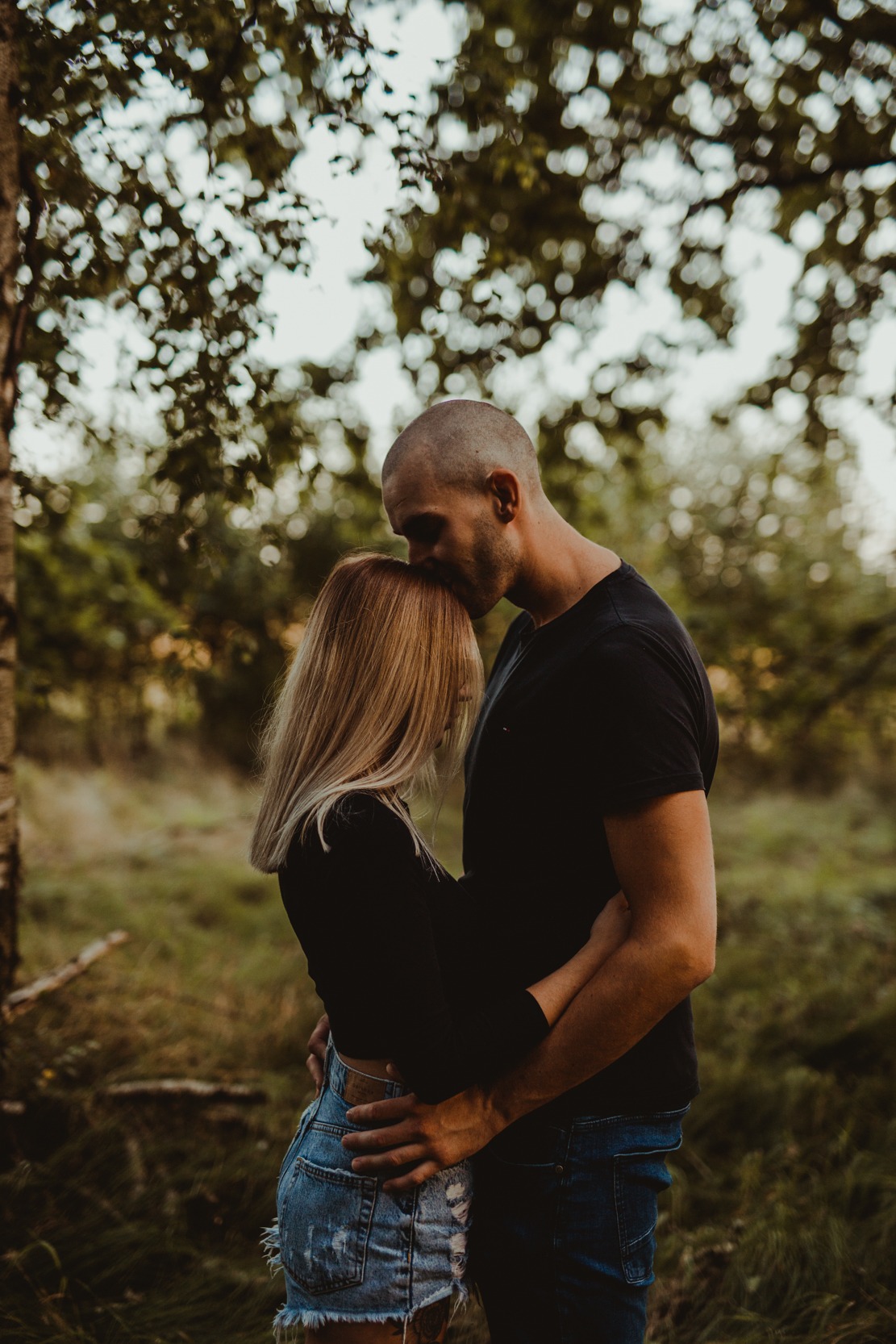 Zakochana para w objęciach na tle leśnej scenerii, mężczyzna całuje kobietę w głowę, ona w krótkich jeansowych spodenkach i czarnej bluzce.