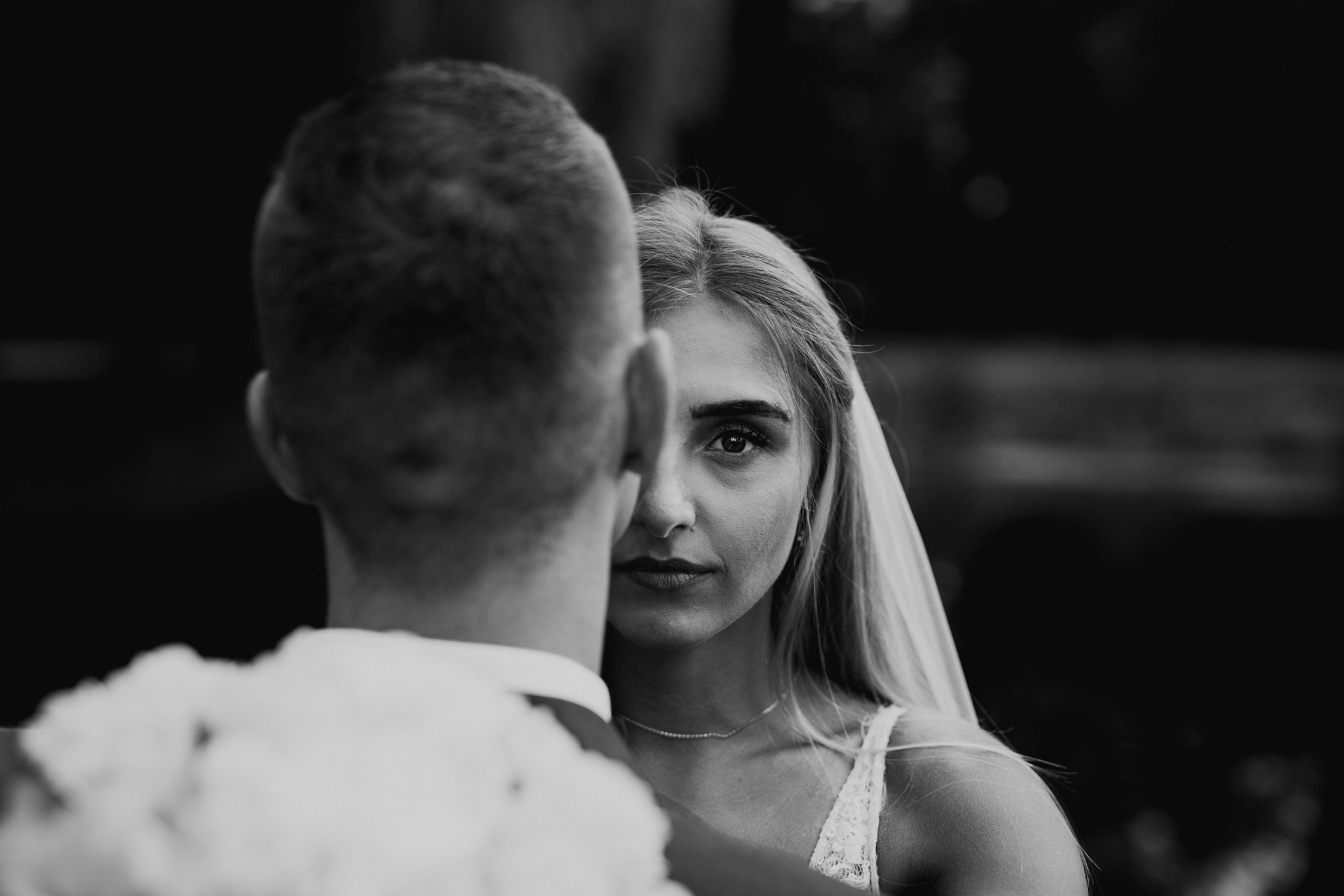 Czarno-białe zbliżenie na twarz panny młodej z welonem, spoglądającej w obiektyw, z widoczną częścią sylwetki pana młodego od tyłu.