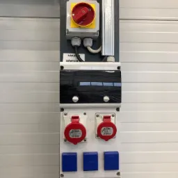 Panel sterowania z wyłącznikiem głównym z czerwoną dźwignią i gniazdami przemysłowymi SCAME, zamontowany na ścianie z białych paneli.