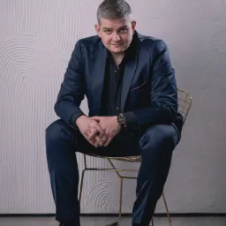 Mężczyzna w granatowym garniturze i czarnej koszuli siedzi na złotym krześle na tle ściany z dekoracyjnym wzorem.