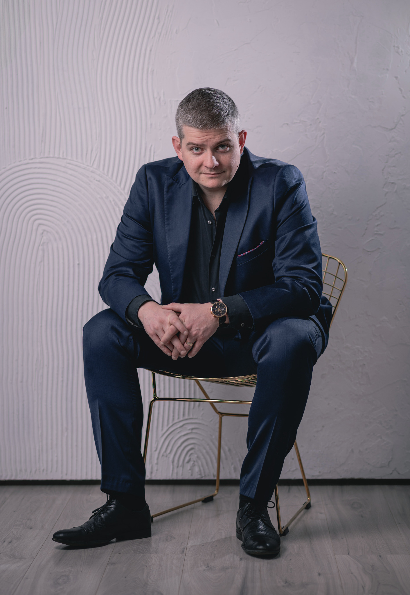 Mężczyzna w granatowym garniturze i czarnej koszuli siedzi na złotym krześle na tle ściany z dekoracyjnym wzorem.