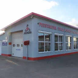 Budynek stacji kontroli pojazdów z szarymi ścianami, czerwonym dachem i napisem 'PODSTAWOWA STACJA KONTROLI POJAZDÓW' nad oknami, logo 'AKROMA' z boku i bramą wjazdową z napisem 'WJAZD'.