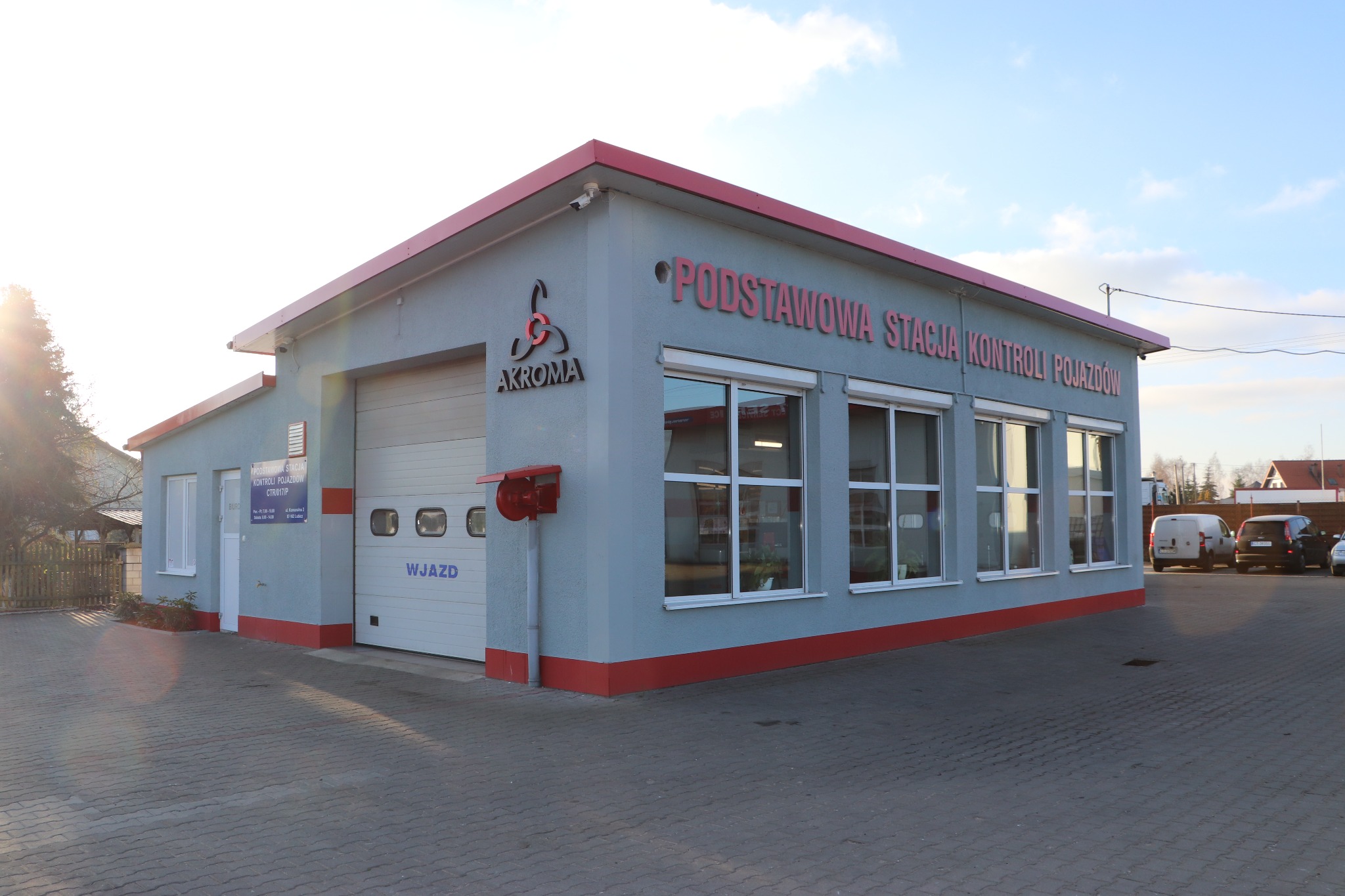 Budynek stacji kontroli pojazdów z szarymi ścianami, czerwonym dachem i napisem 'PODSTAWOWA STACJA KONTROLI POJAZDÓW' nad oknami, logo 'AKROMA' z boku i bramą wjazdową z napisem 'WJAZD'.