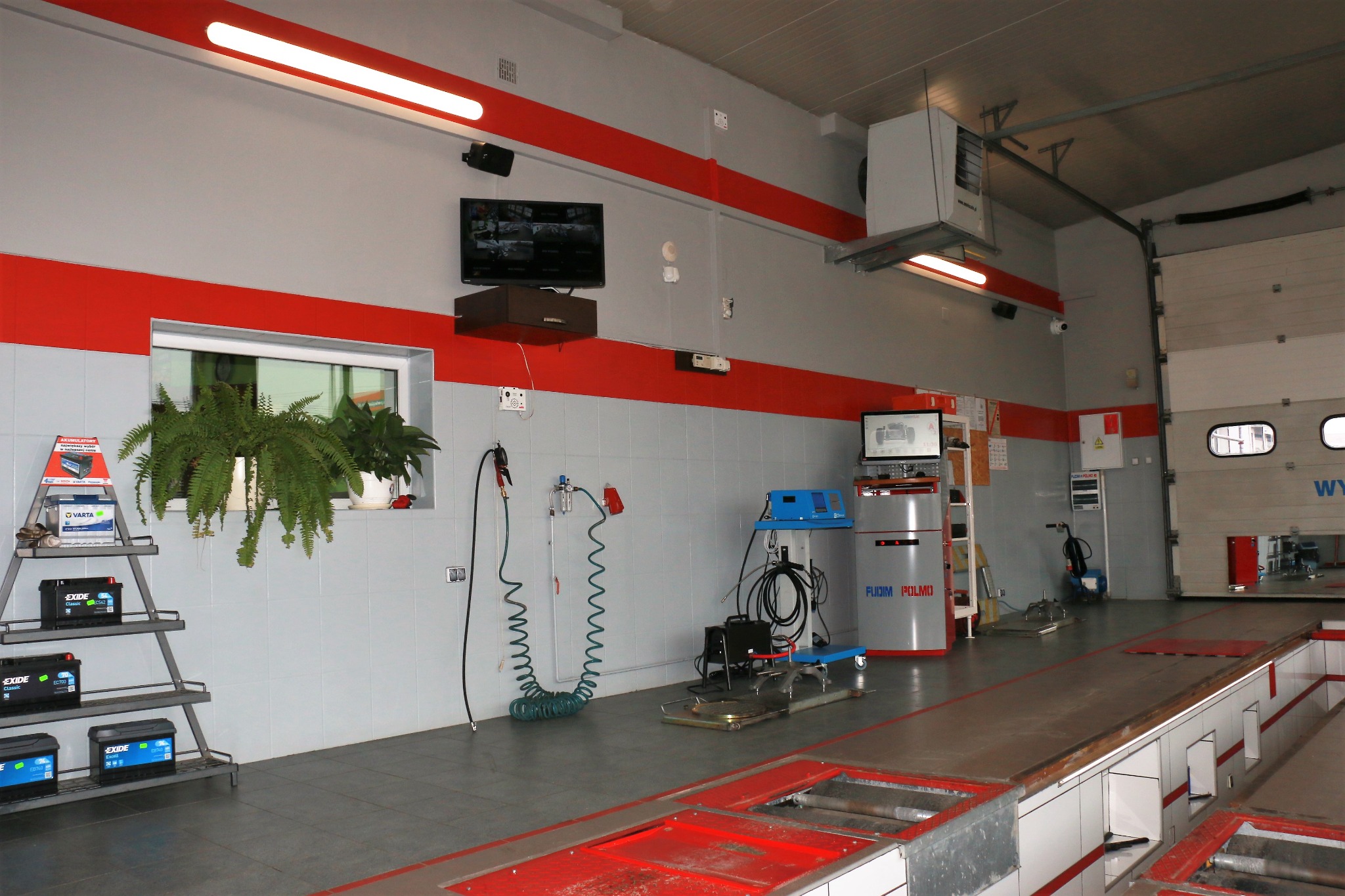 Wnętrze warsztatu samochodowego z podnośnikiem najazdowym, testerem akumulatorów na regale przy oknie i sprzętem diagnostycznym, ściany w kolorach szaro-czerwonych.