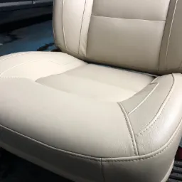 Beżowe skórzane siedzenie samochodowe z wyraźnym szwem, sfotografowane z bliska na tle ciemnej podłogi.