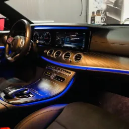 Wnętrze luksusowego samochodu Mercedes-Benz z podświetleniem LED w kolorze niebieskim, widoczny kokpit z drewnianym wykończeniem, wyświetlacz multimedialny i skórzane fotele. Na desce rozdzielczej...