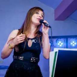 Kobieta w czarnej sukience z mikrofonem śpiewa na scenie, oświetlona niebieskim światłem, z widocznym złotym zegarkiem na nadgarstku.