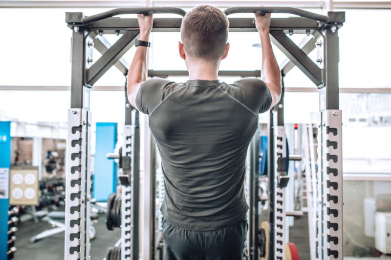Mężczyzna wykonuje podciąganie na drążku w siłowni, widok z tyłu, z widocznym sprzętem do ćwiczeń w tle.
