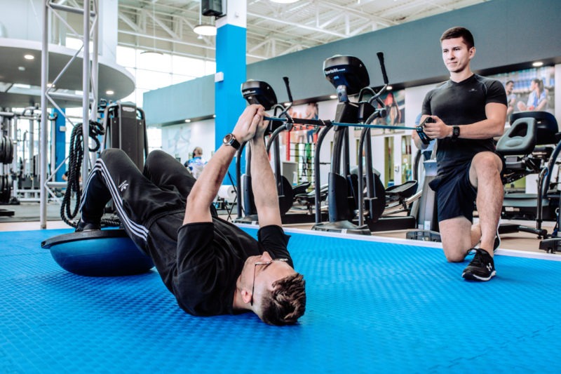 Mężczyzna w czarnym dresie Adidas leży na niebieskiej półkuli Bosu Ball, trzymając gumę do ćwiczeń, podczas gdy drugi mężczyzna w czarnym stroju sportowym klęczy obok, trzymając drugi koniec gumy...