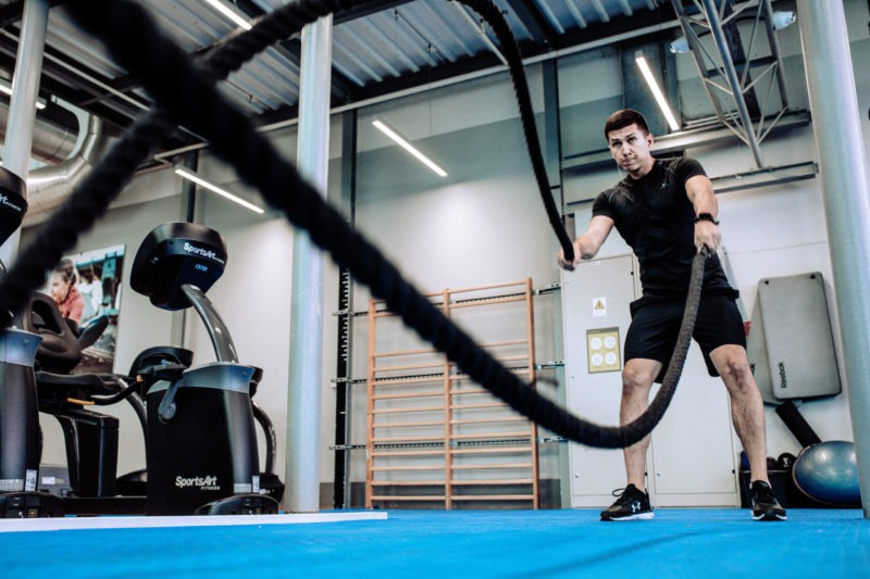 Mężczyzna w czarnym stroju ćwiczy dynamicznie z grubymi linami w siłowni, w tle sprzęt do ćwiczeń i drabinki gimnastyczne.