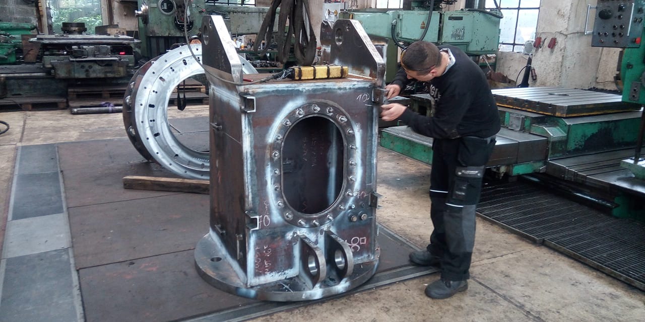 Pracownik w okularach ochronnych dokonuje pomiarów metalowej konstrukcji z owalnym otworem, widoczna spawana obręcz, w tle maszyny przemysłowe.