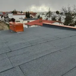 Szary, płaski dach pokryty papą termozgrzewalną z widocznym kominem wentylacyjnym w kolorze pomarańczowym na tle zabudowy miejskiej i pochmurnego nieba.