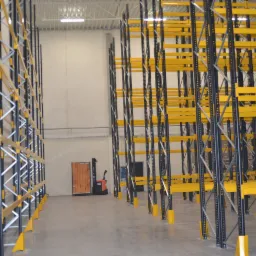 Wnętrze magazynu z rzędami wysokich, czarno-żółtych regałów metalowych; w tle widoczny wózek widłowy i palety.