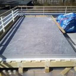 SW Projekt - Płyta warstwowa na dachu budynku, zabezpieczona drewnianą konstrukcją i folią, z widocznymi budynkami w tle.