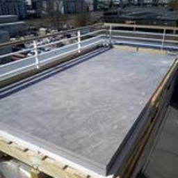 SW Projekt - Płaski dach z szarej płyty warstwowej, zabezpieczony białą barierką, widok z góry na tle miejskiej zabudowy w słoneczny dzień.