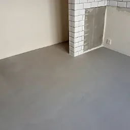 Szara, wylewana podłoga w rogu pomieszczenia z fragmentem ściany wyłożonej białymi, prostokątnymi płytkami, częściowo bez fugi i z widocznym gniazdkiem elektrycznym.