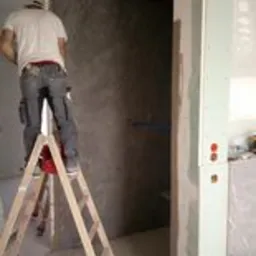 Pracownik na drewnianej drabinie montuje izolację na ścianie wewnątrz budynku, widoczne profile sufitowe i instalacja elektryczna