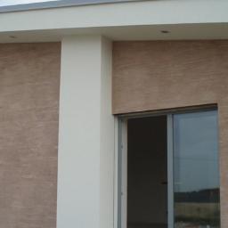 SW Projekt - Fragment elewacji budynku z oknem i przesuwnymi drzwiami, wykończony tynkiem o fakturze imitującej drewno, z widocznym samochodem dostawczym w tle.