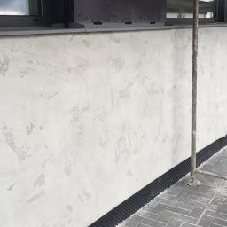 SW Projekt - Jasna elewacja budynku z dekoracyjną fakturą, widoczna czarna membrana izolacyjna przy gruncie i fragment rusztowania na chodniku z kostki.