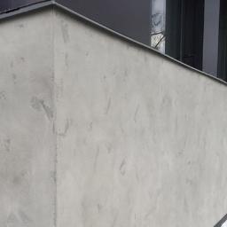 SW Projekt - Ściana budynku z szarą, dekoracyjną elewacją o nieregularnej fakturze, widoczne czarne okna i worki z materiałem budowlanym w tle.