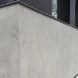 Ściana budynku z szarą, dekoracyjną elewacją o nieregularnej fakturze, widoczne czarne okna i worki z materiałem budowlanym w tle.