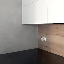 SW Projekt - Fragment kuchni po remoncie: szara ściana z trzema gniazdkami w chromowanej ramce, drewniana okładzina ściany nad blatem z dwoma gniazdkami i białe szafki wiszące.