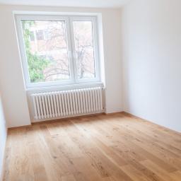 SW Projekt - Pusty pokój po remoncie z drewnianą podłogą, białymi ścianami i grzejnikiem pod oknem z widokiem na drzewa.