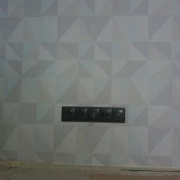 SW Projekt - Ściana pokryta tapetą w geometryczne, trójwymiarowe wzory w odcieniach szarości, z zamontowanym czarnym panelem gniazdek i włączników.